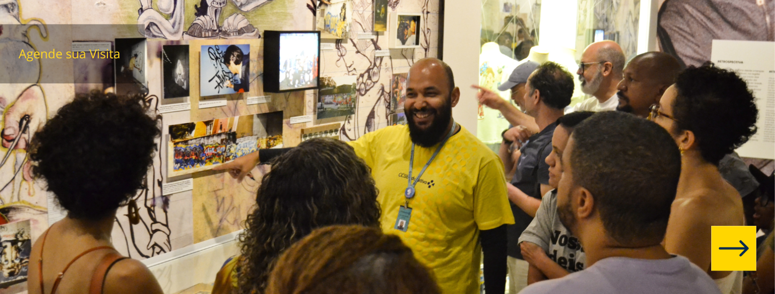 Educador aponta para obra na sala de exposições enquanto é observado por diversos visitantes.